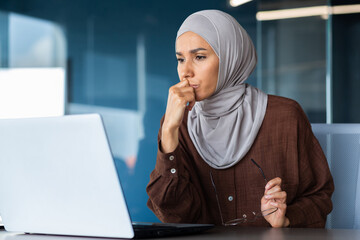 Portrait of a serious young woman in a hijab sitting in front of a laptop, holding glasses,...