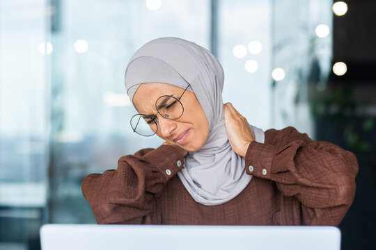 Close-up photo. Tired young arab woman in hijab student studying at laptop and holding neck, wincing in pain, massaging with hands.