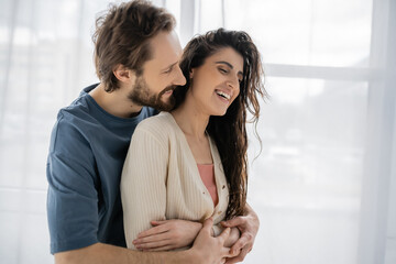 Bearded man embracing overjoyed brunette girlfriend at home.