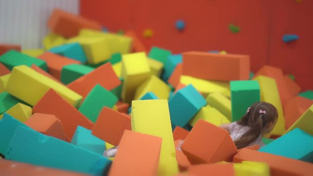 Little happy girl play and have fun in a dry pool with soft multicolored cubes in a children's entertainment center and jump on the paralon blockes in the playroom.