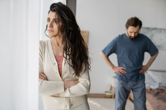 Displeased woman standing near curtains during relationship crisis with blurred boyfriend at home.