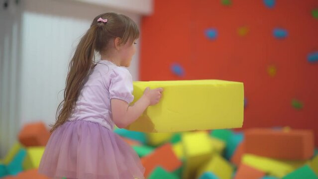 Little happy girl play and have fun in a dry pool with soft multicolored cubes in a children's entertainment center and throw paralon block in the playroom.