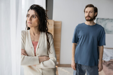Offended woman standing near curtain while blurred boyfriend talking at home.