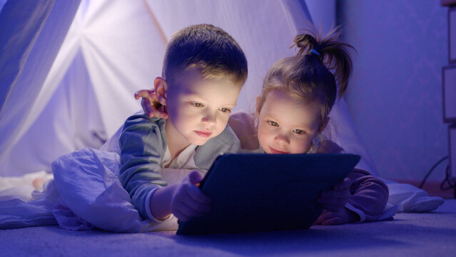 Children lie in game house embracing and laugh watching cartoon on tablet with interest