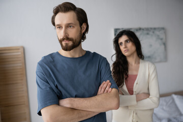 Sad man crossing arms near blurred girlfriend in bedroom at home.
