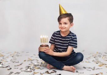A boy of 7 years old in a striped T-shirt and jeans in a gold cap celebrates his birthday. He holds...