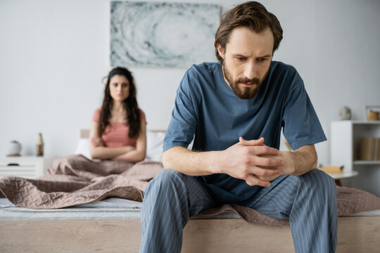 Tensed man in pajama sitting near blurred girlfriend in bedroom at home. - Powered by Adobe