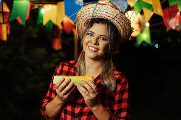 Brazilian woman wearing typical clothes for the Festa Junina - June festival - eating corn on the...