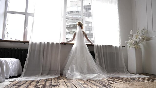 the bride stands at the window in a luxurious wedding dress
