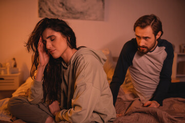 Upset woman sitting near blurred boyfriend talking on bed in evening.