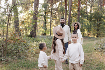 Fototapeta premium Young mother, father with daughter and sons are walking, having fun in autumn forest. Family holding hands enjoying time together at background of trees. Happy parenthood and childhood concept.