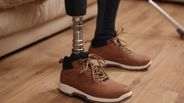 sofa sitting, assistive device, independent living. Close-up shows man with prosthetic leg putting on shoes on sofa, crutches resting beside him.
