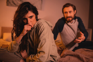 Frustrated woman sitting near boyfriend quarreling in bedroom in evening.