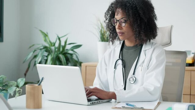 Video of confident female doctor using her mobile phone while working on laptop in the consultation.