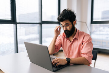 Thoughtful Indian businessman wearing casual clothes working on laptop sitting at table in light office room on background of window. Bearded freelancer male in glasses looking at computer screen.
