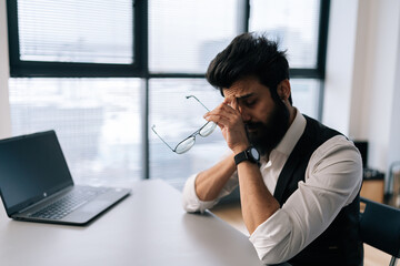 Side view of tired exhausted Indian businessman massaging nose bridge, taking off glasses, feeling eyestrain, sitting at work desk. Overworked freelancer male suffering from dry eye syndrome.