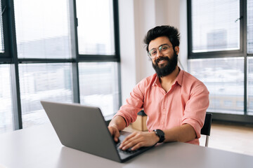 Cheerful Indian business man in eyeglasses sitting at desk with laptop smiling looking at camera in light coworking office by window. Bearded freelancer male posing working on notebook alone.