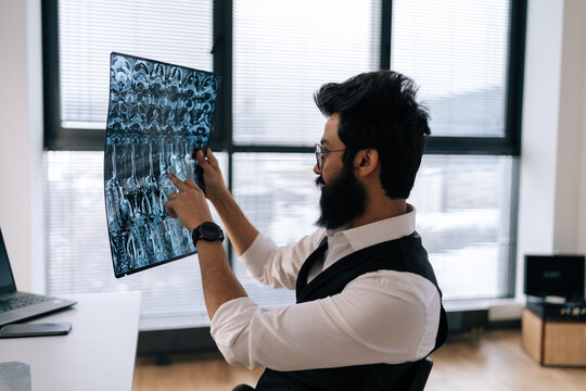Side view of professional Indian male doctor in glasses thinking on diagnosis looking at MRI images scan by window. Radiology clinic physician in stylish clothes examining spinal column x-ray image.