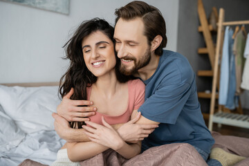 Overjoyed man hugging brunette girlfriend in pajama on bed at home.
