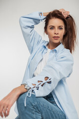Studio fashion female portrait of a beautiful young woman in stylish casual denim clothes with a blue shirt and jeans sits and poses on a white background