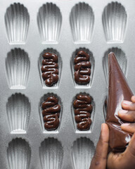Piping chocolate Madeleine batter into a greased Madeleine pan, chocolate cake batter in a baking pan