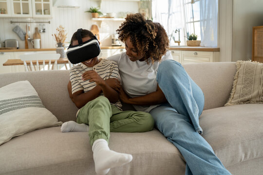 Happy african american mom and son using virtual glasses for play video games. Boy looking at virtual world in VR helmet sitting on couch at home. Virtual reality, cyberspace, entertainment concept.