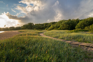 Summer rural landscape with green field, path and lake at sunset. Natural scenery in village, cloudy day in countryside. Recreation in nature concept
