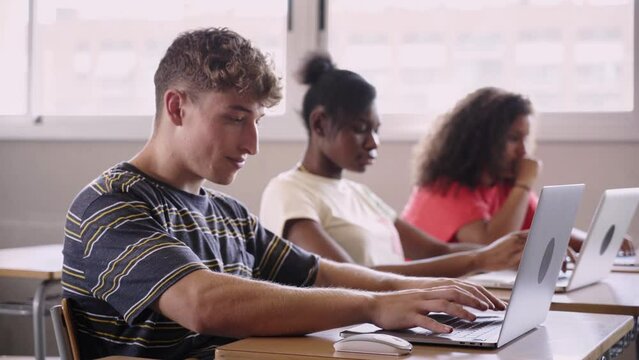 Multi-ethnic group of three young high school students study together using computers. Digitized education and technology. Focus on the boy typing on laptop. Generation z people.