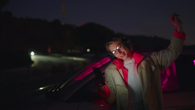 Caucasian female enjoys carefree night as she jumps near car and happily dances with favorite pop music in headphones. Slow motion, attractive smiling hipster girl listen online music via mobile app
