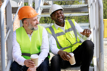 Smiling multicultural builders in hardhats, holding coffee sitting on stairs at construction site....