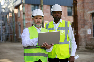 Portrait ofthe two builders together standing at construction area with wireless laptop working on...