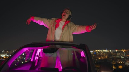 Happy and satisfied female hums favorite song while listening online music on headphones. Dancing young woman enjoying playlist on mobile phone application while standing in convertible car at night
