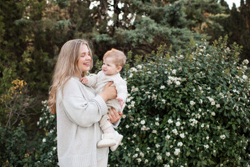 Happy smiling young mother holding baby 1 year old wearing stylish casual knitted clothes standing over blooming bushes with flowers in park outdoor. Motherhood. Maternity.