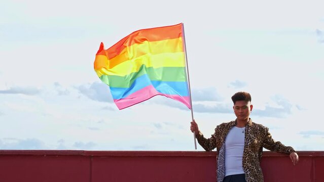 young gender non binary asian person from the front holding a gay pride flag blowing in the wind