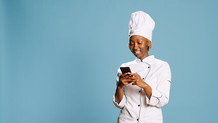 Smiling cheerful chef browsing online social media page on camera, feeling happy scrolling through internet website. Gourmet cook in gastronomy uniform using mobile phone on blue backdrop.