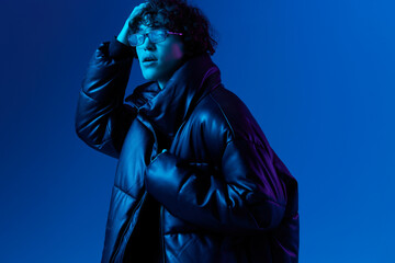 Obraz premium Man in winter down jacket model posing with glasses, hipster lifestyle, portrait blue background, mixed neon light, fashion style and trends guys teenager, copy space