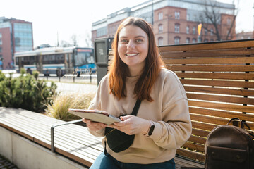 Beautiful smiling female student using tablet outdoors. Happy cheerful young woman siting on the bench on city street, Urban lifestyle concept. Online education or shopping banner