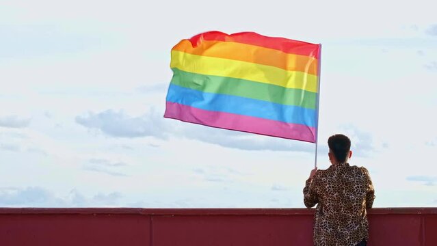 young gender non binary asian person with back to back holding a gay pride flag blowing in the wind