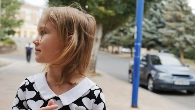 A cute caucasoid girl with blond hair is dressed in a shirt with drawings of hearts walking along the sidewalk and looking in different directions turning her head. teenager walking the streets alone