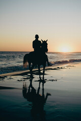 Silhouette of a rider on his horse walking on the seashore with the sunset of a summer afternoon on the beach in the background
