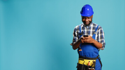 Construction worker texting messages on smartphone app, using social media browser in studio. Young...