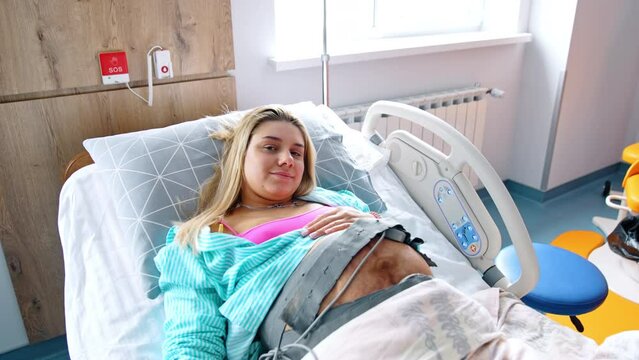 Blonde pregnant woman lies in bed with cardiotocography belt on the belly. Lady smiles to the camera. Top view.