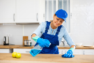 Positive skilled female janitor in blue uniform and rubber gloves cleaning worktops at kitchen