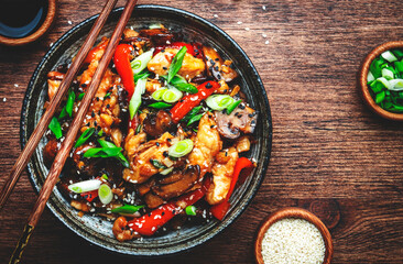 Stir fry chicken with paprika, mushrooms, green chives and sesame seeds in ceramic bowl.  Asian cuisine dish. Wooden kitchen table background, top view