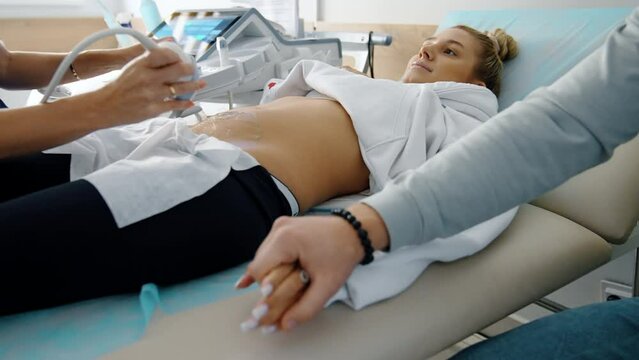 Beautiful pregnant lady undergoing ultrasound examination. Male hand takes his wife’s hand showing love and support.