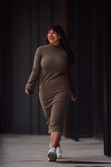 A confident and stylish plus-size woman walking by a modern building in an olive green dress,...