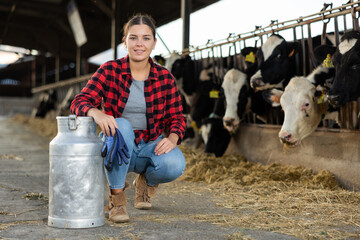 Positive young woman farmer squatting at milk can in cowshed and smiling.