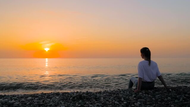 Slow motion: portrait of woman in white t-shirt is sitting on the sea shore and looking at the warm sunset sky. Sea water waves are crashing on beach. Freedom, calm, seascape and summer concept