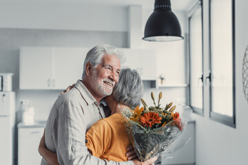 Old man giving flowers at his wife sitting on the sofa at home for the San Valentines’ day....
