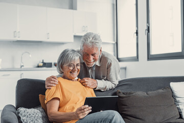 Happy older couple using computer tablet together at home, excited mature man and woman looking at mobile device screen, shopping or chatting online, sitting on cozy couch in living room. © Daniel